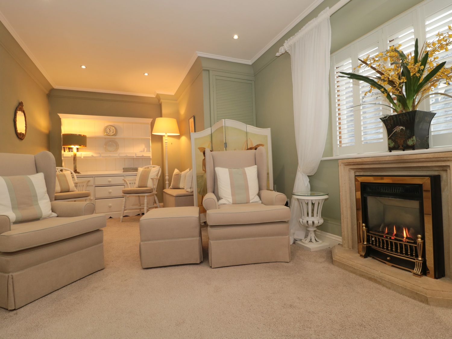 A living room with beige armchairs and ottomans a fireplace with flowers on the mantel and a white cabinet with chairs at Tree Tops in Broadway