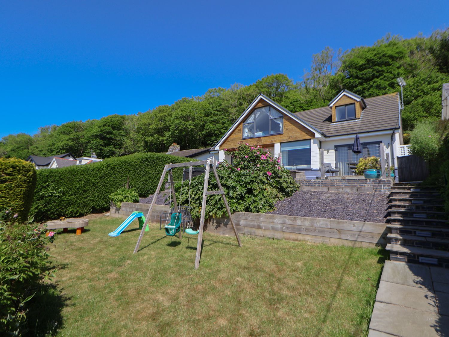 A backyard with a swing set slide and garden toys in front of a house with a patio and stairs at Buzzards View in Dyserth