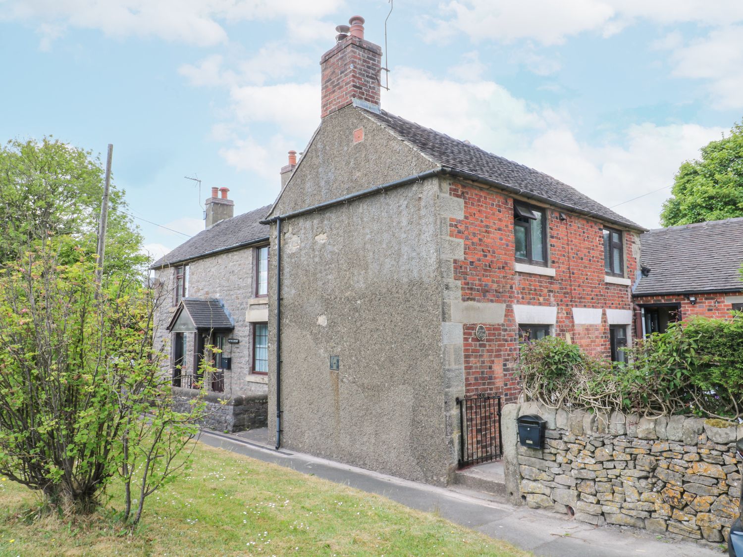A brick and stone house with a small gated entrance and a stone wall surrounded by grass and trees at Corner Cottage in Ashbourne