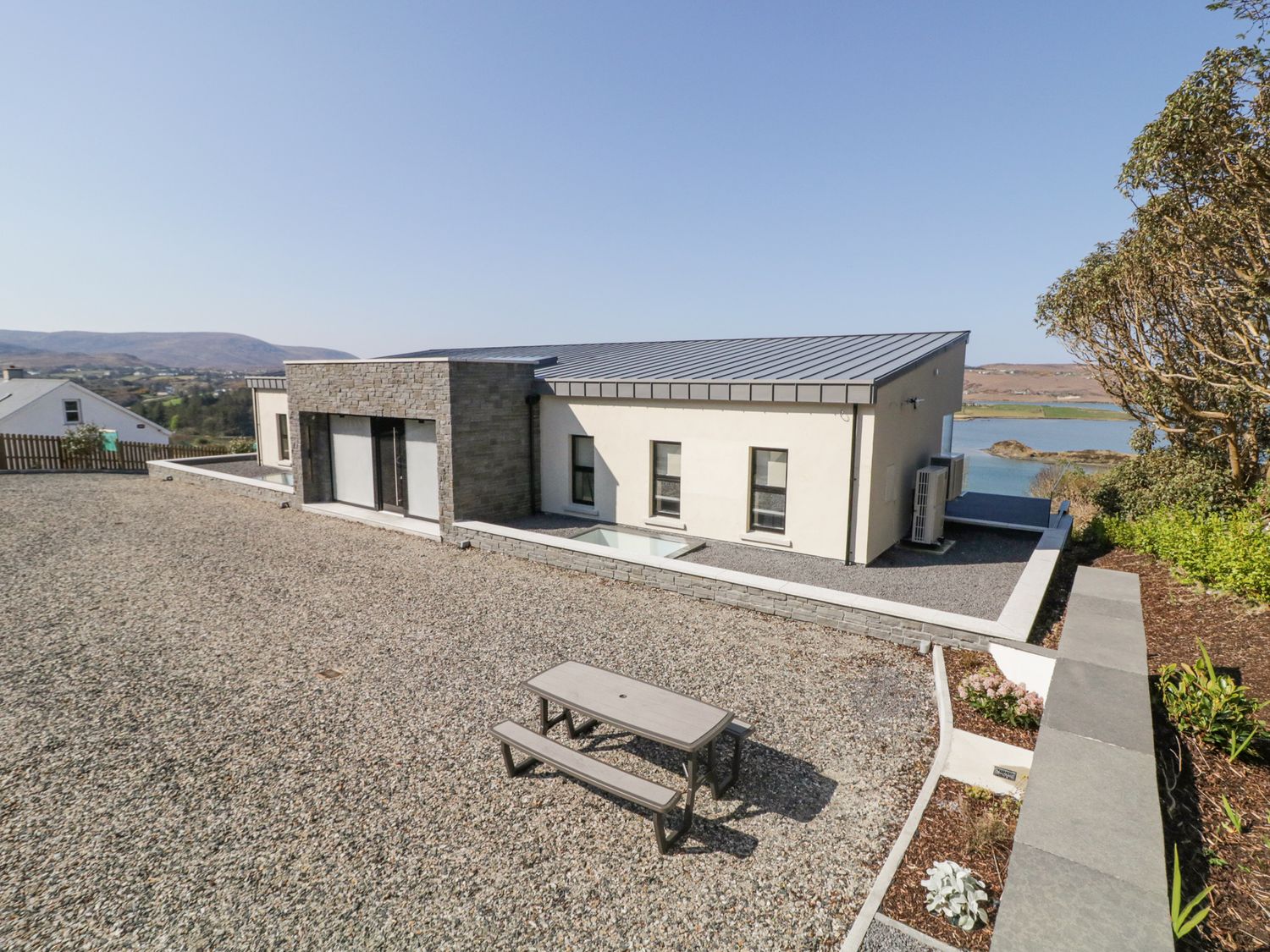 A modern one-story house with a gravel yard and a picnic table near a body of water at Atlantic Sea Breeze Near Gortahork County Donegal