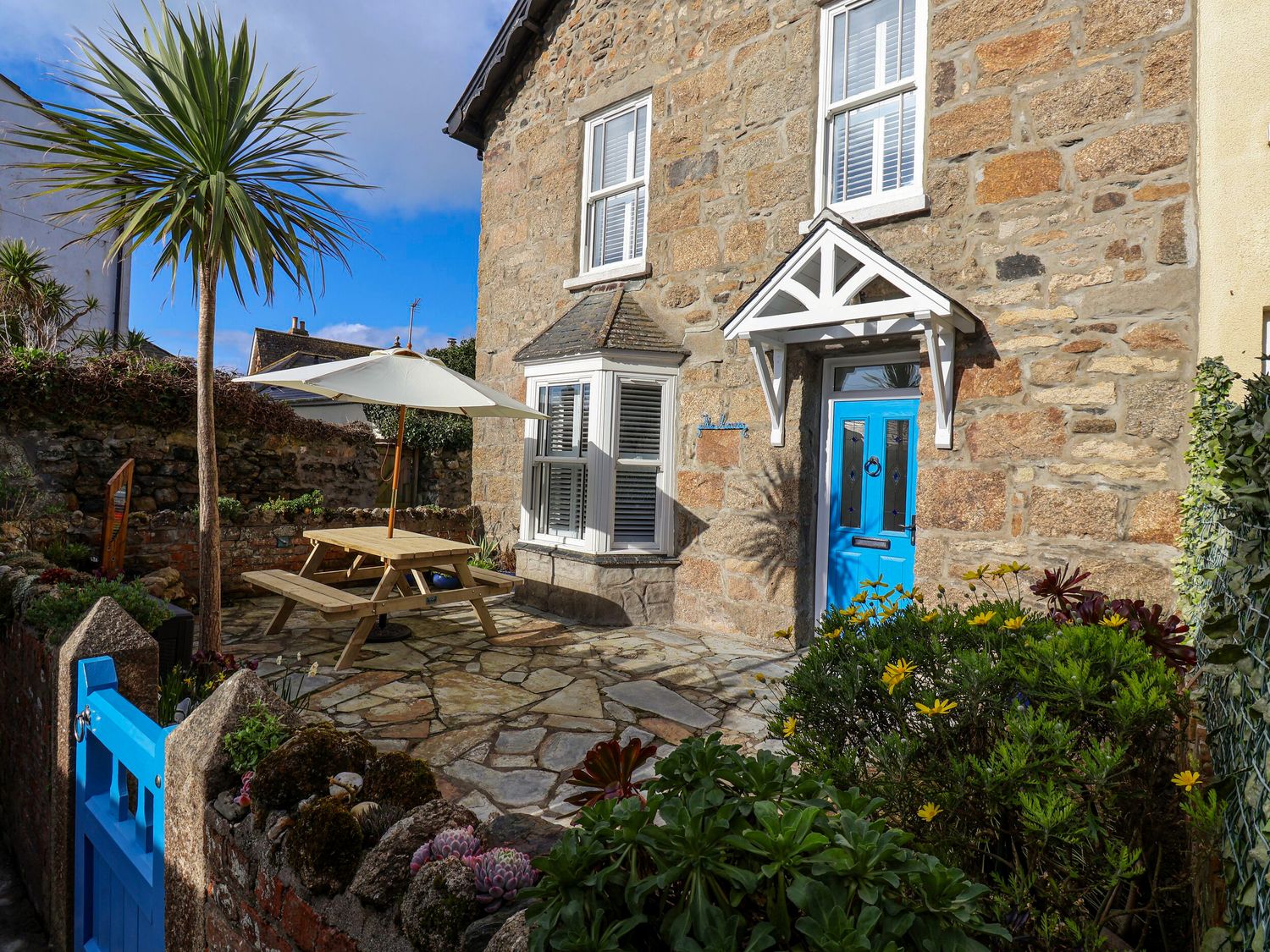 A stone house with a blue door and window frames a picnic table with umbrella and garden plants at The Haven in Marazion