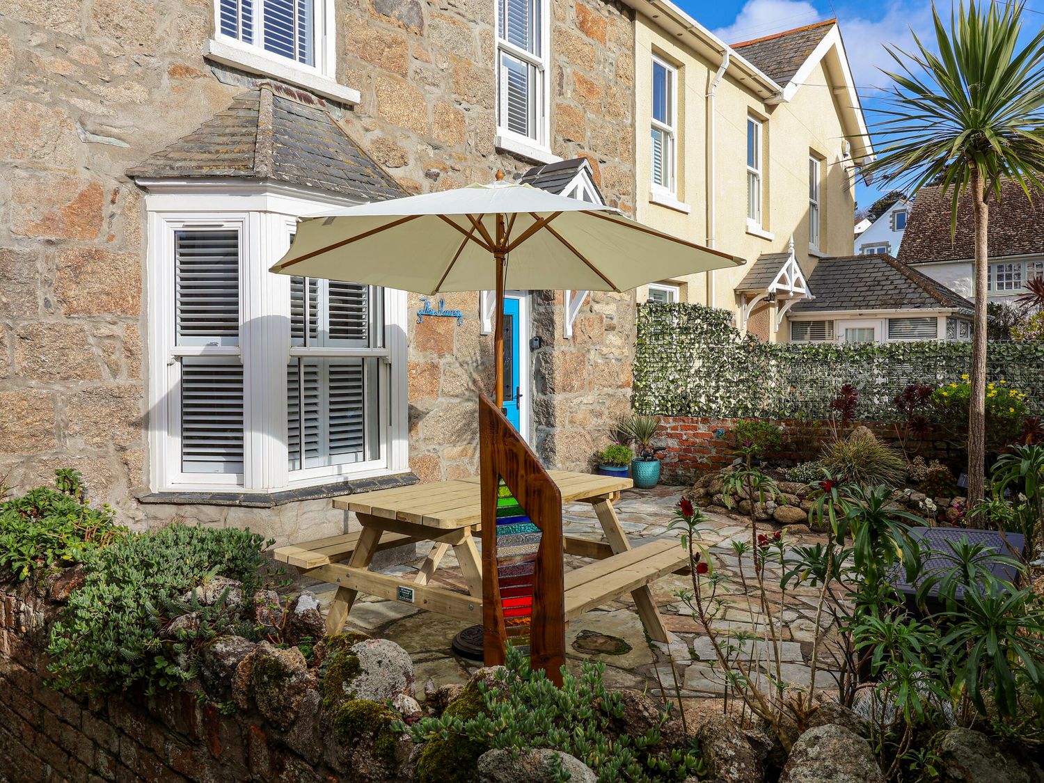 A stone house with a wooden picnic table and umbrella in a garden with plants and a stone patio at The Haven in Marazion