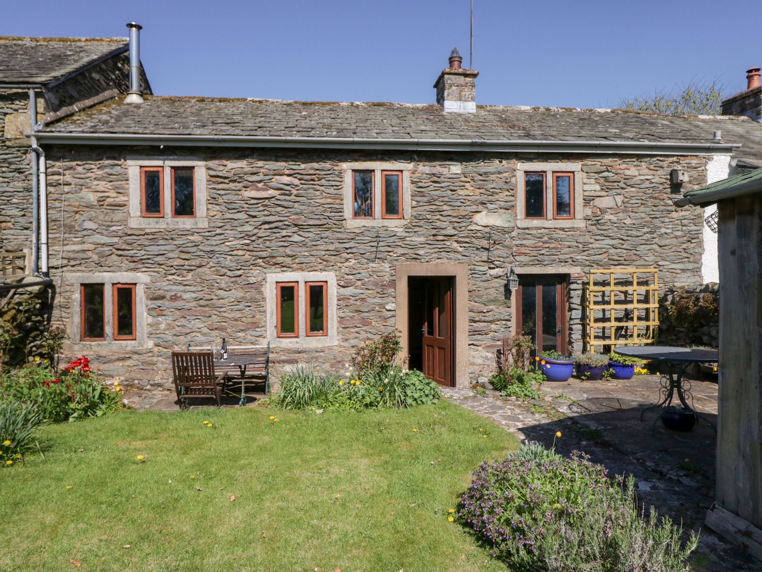 A stone cottage with wooden windows and doors a garden with grass flowers and outdoor furniture at Tethera Cottage in Bampton Cumbria