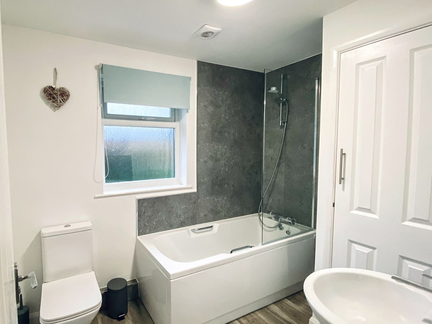 A bathroom with a bathtub shower, toilet, window with blind, sink, and a door at Trelawne Near Crackington Haven