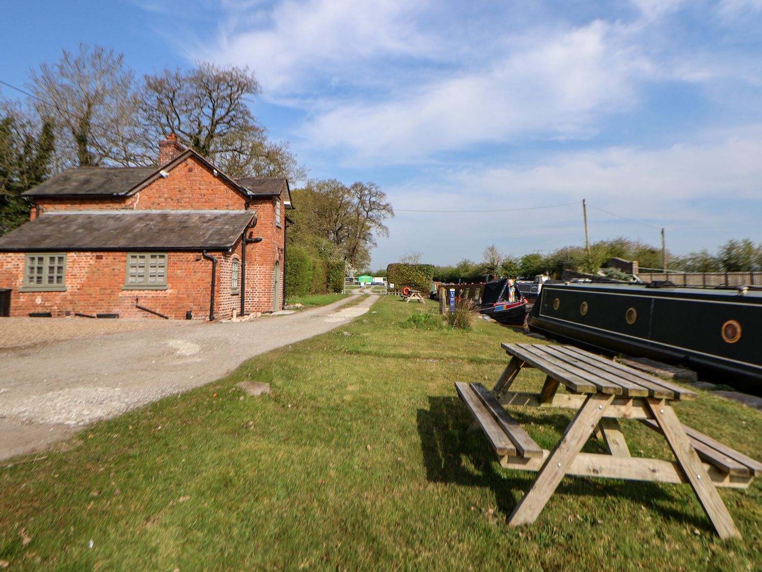 A wooden picnic table on grass near boats and a brick house by a gravel path at Canal Basin Cottage in Nantwich
