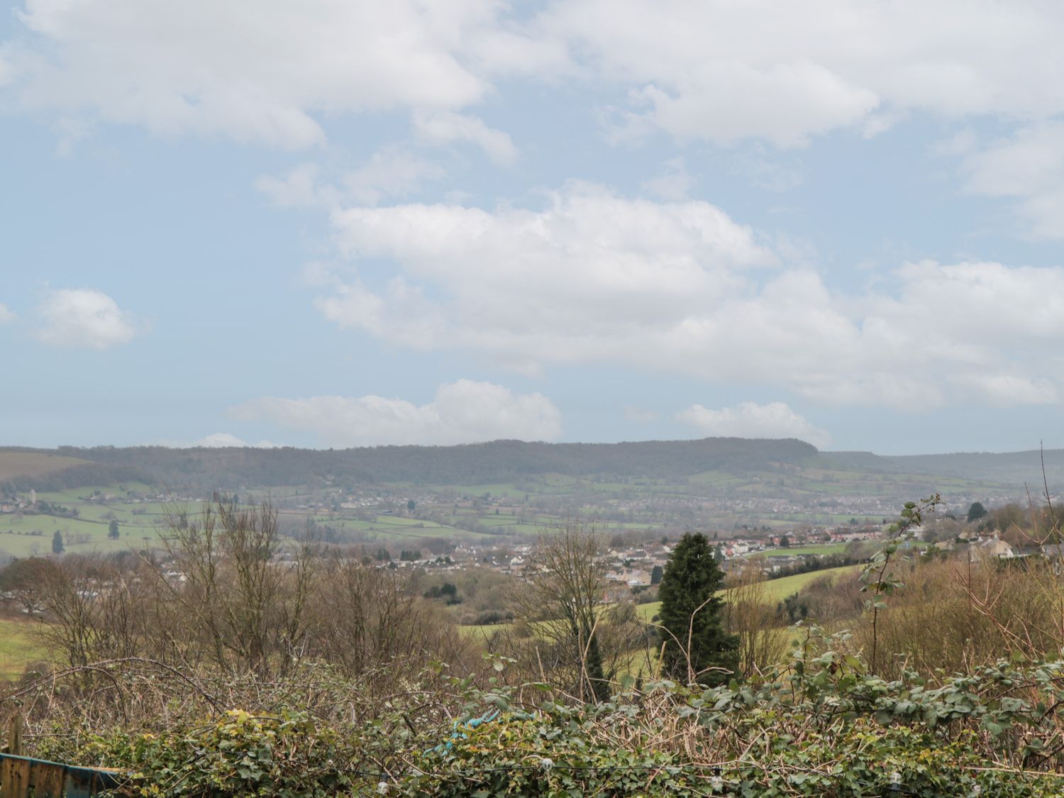 A rural landscape with bushes and trees overlooking a village and hills under a cloudy sky at Twin Room in Whiteshill