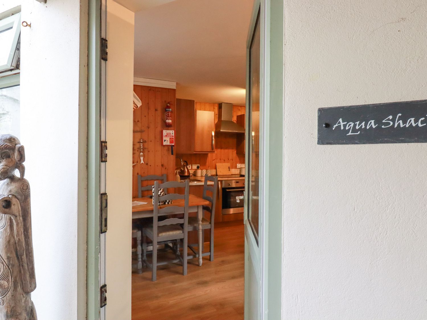 View through an open door into a kitchen dining area with wooden chairs and table and kitchen cabinets at The Aqua Shack in Newquay
