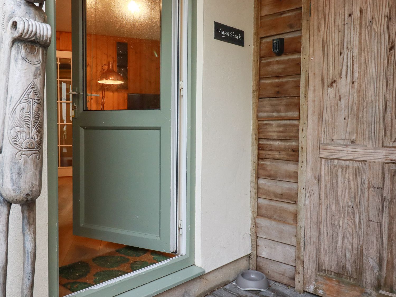 An open green door leading to a room with wooden walls and a lamp a carved wooden sculpture near the door a wooden door and a pet bowl on the floor at The Aqua Shack in Newquay