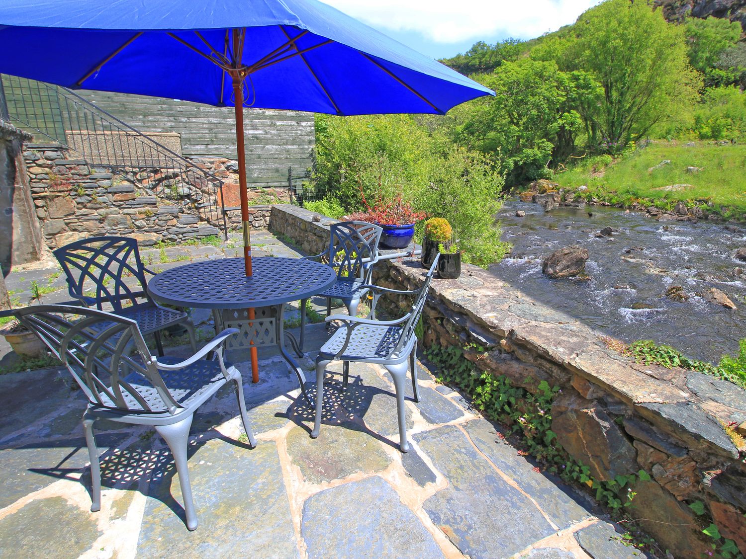 An outdoor patio with metal chairs and a table under a blue umbrella next to a river at Riverbank Cottage in Beddgelert