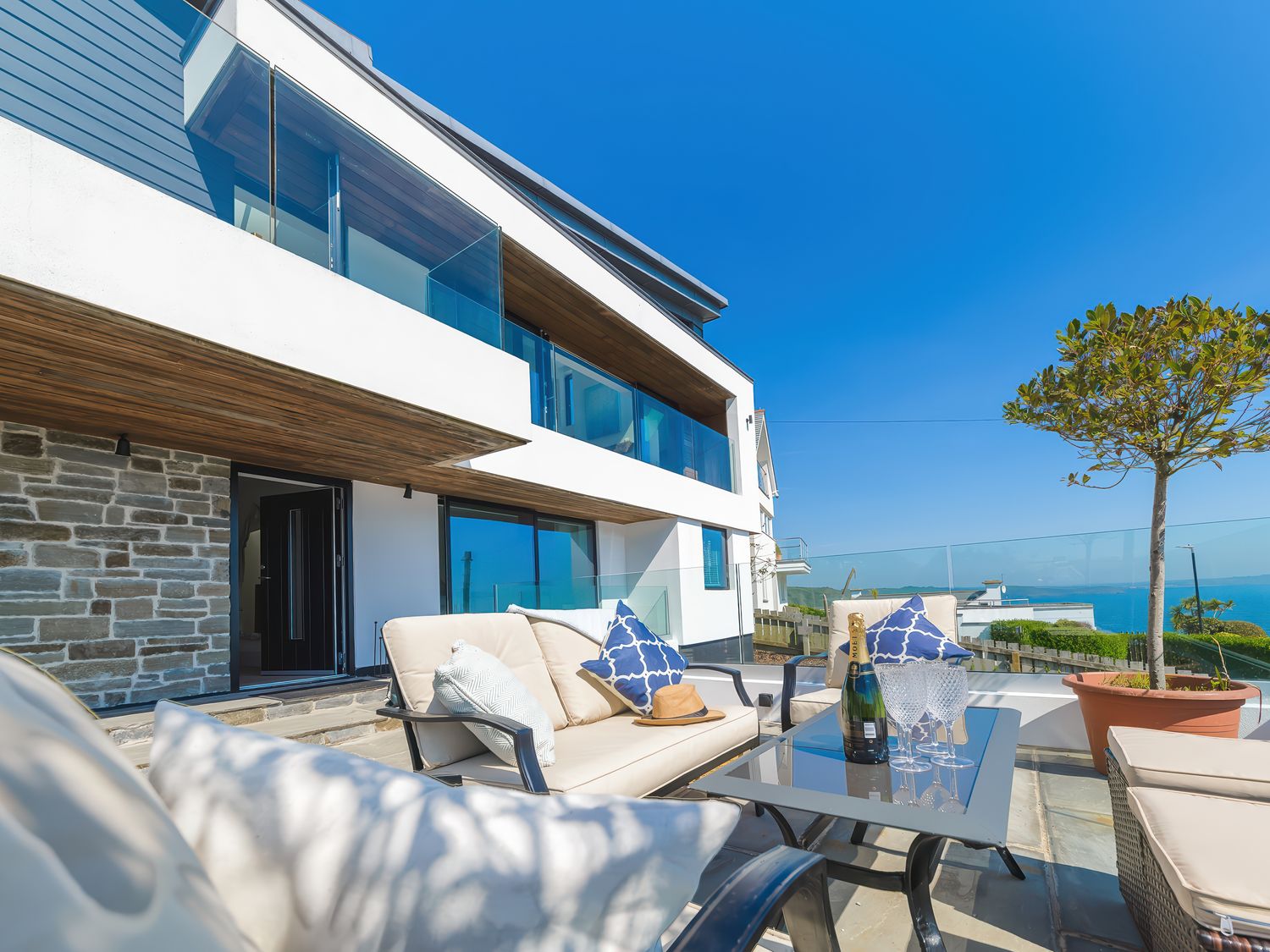 An outdoor patio with cushioned chairs a glass table champagne bottle and glasses a potted tree and a modern house with glass balconies at Oystercatcher in Mevagissey