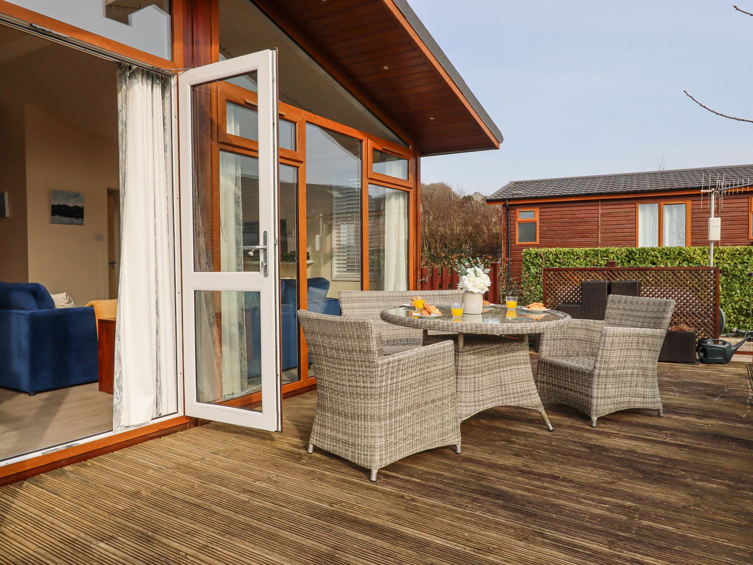 A patio with wicker table and chairs set with drinks and pastries outside a house with glass doors at Matilda Lodge in Newton Ferrers