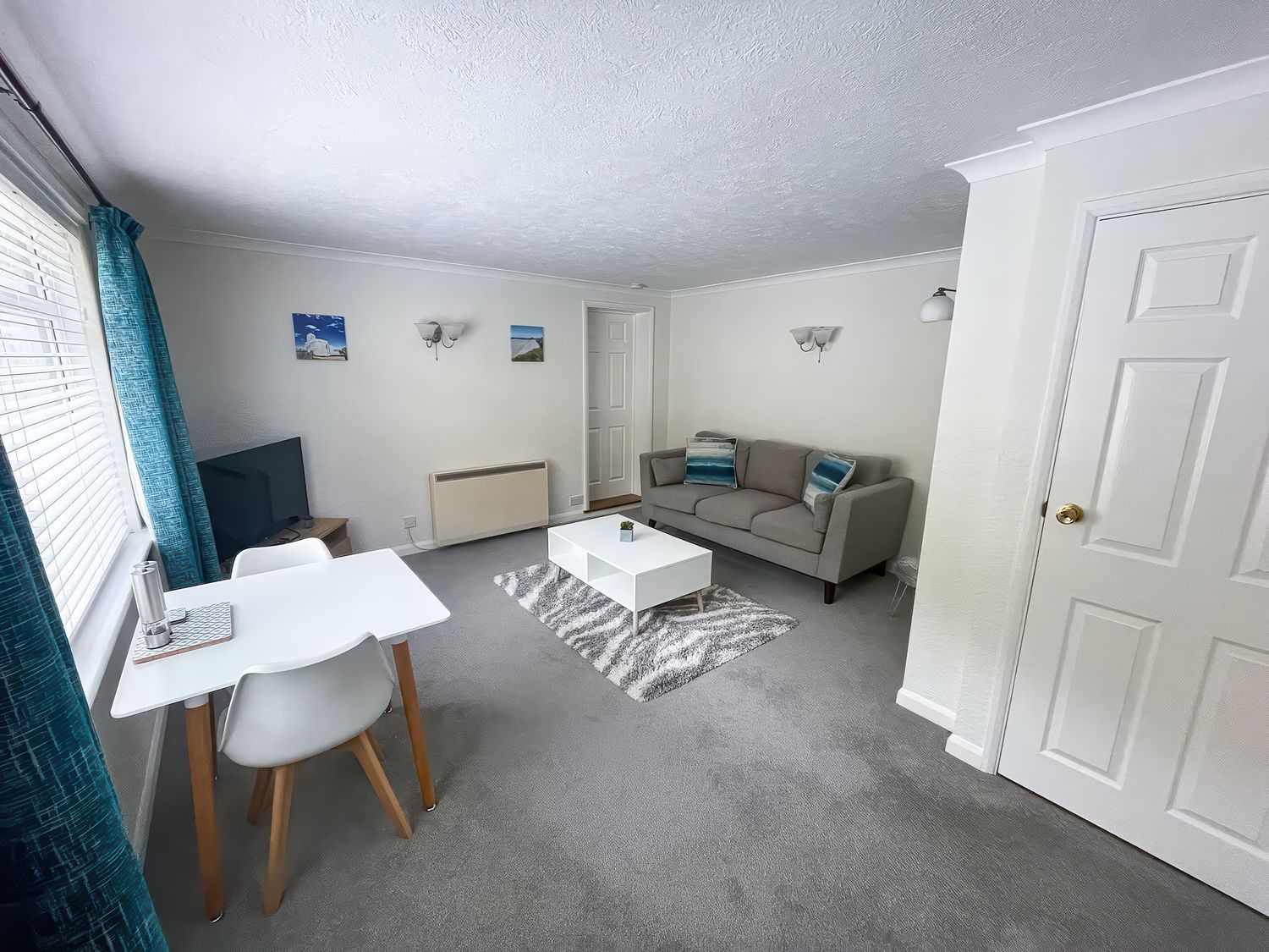 A living room with a gray sofa a white coffee table a dining table with two chairs a television and blue curtains at 2 Chatsworth Riverscape Apartments in Newquay