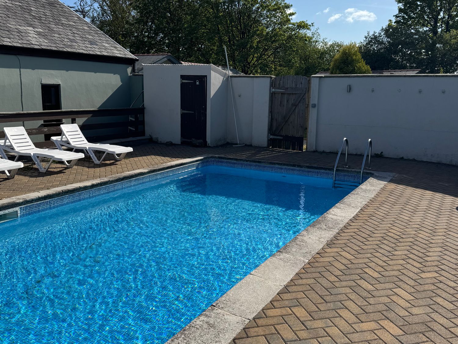An outdoor swimming pool with three white lounge chairs on a paved area at Tig Bhan in Llanteg