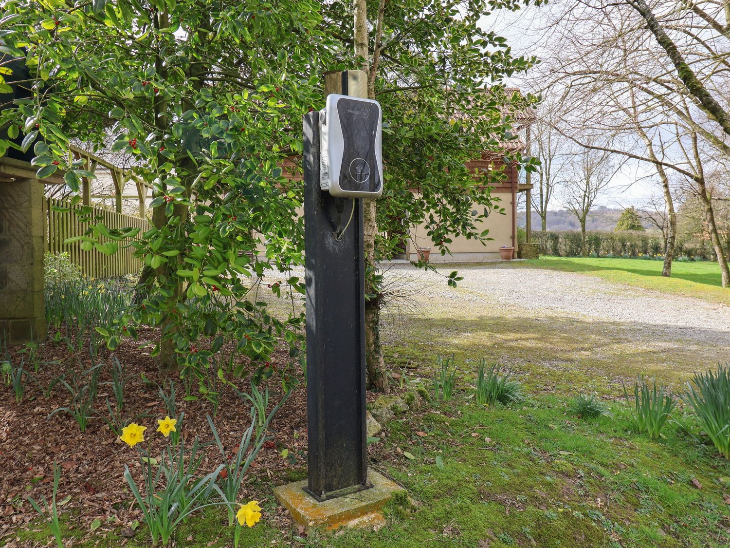 A black electric vehicle charger mounted on a post near grass shrubs and daffodils outside a house in a rural area at Meadow Cottage in Upottery