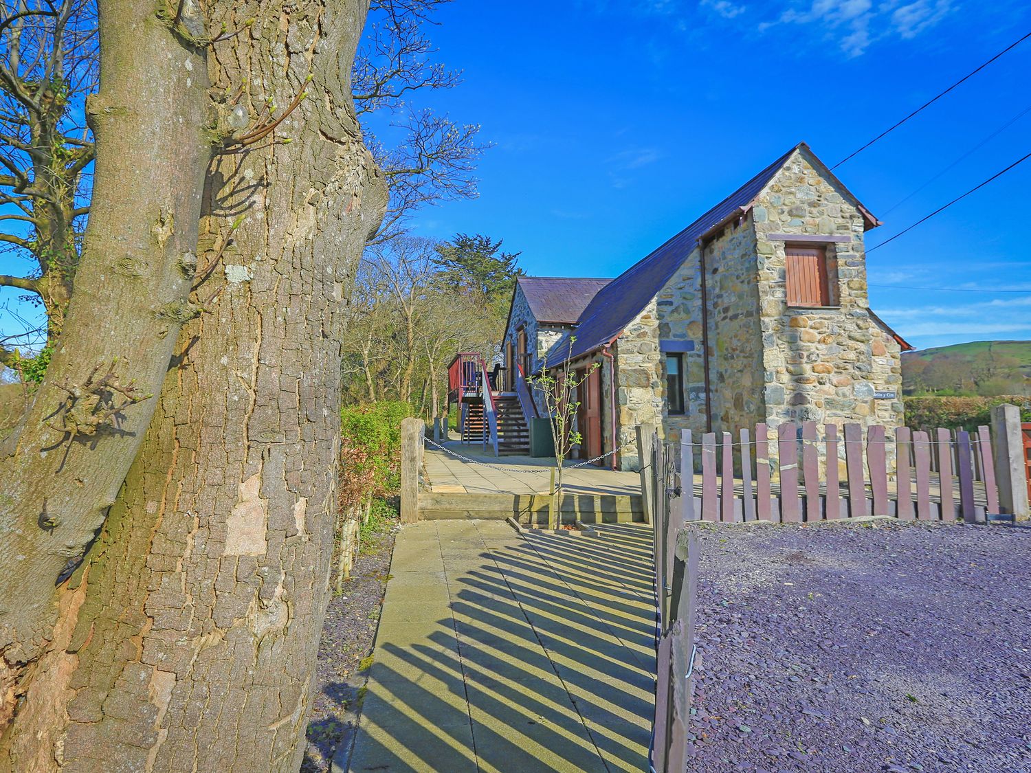 A stone house with a wooden fence and a large tree in front at Melin Y Cim in Caernarfon