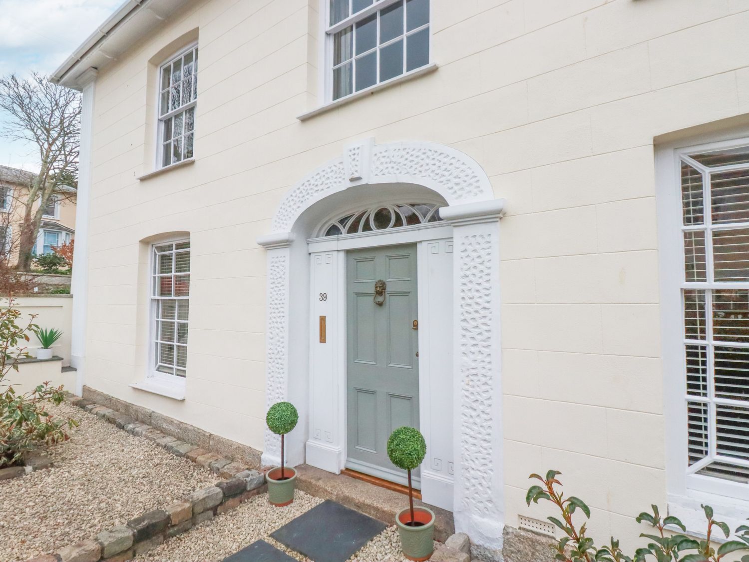 The exterior front entrance of a house with a light green door flanked by two potted topiary plants at Rhondda House in Truro