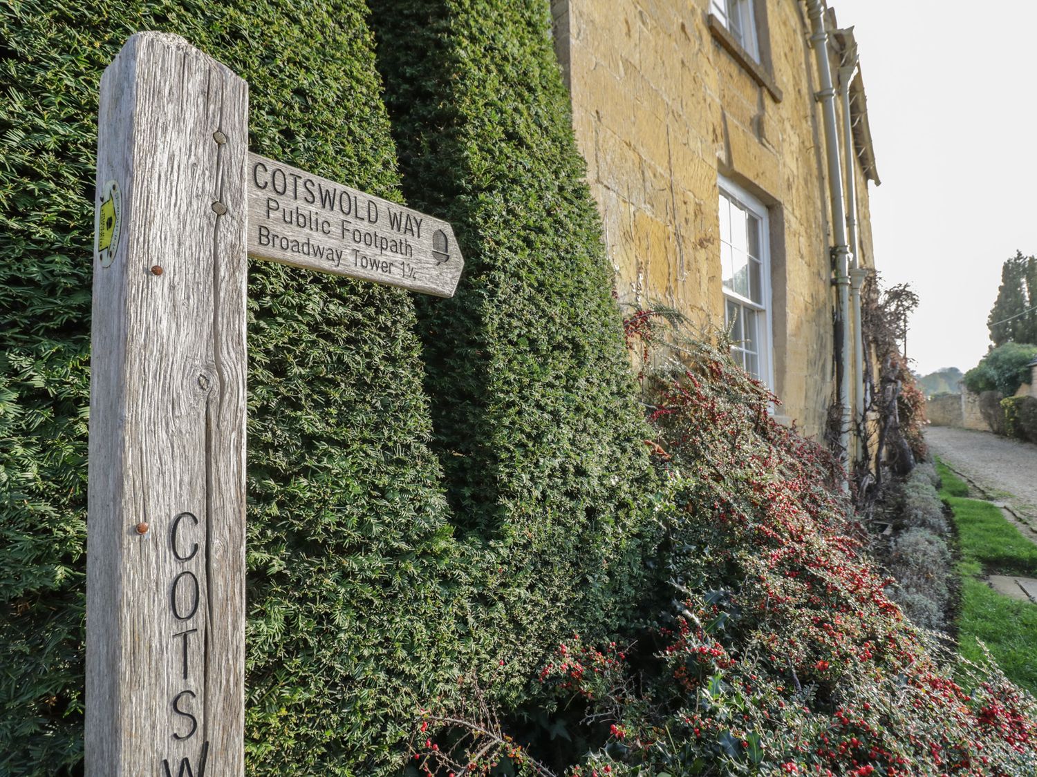 A wooden signpost for Cotswold Way public footpath near a stone building with green hedges and red berries at 2 Summers Way Moreton-In-Marsh