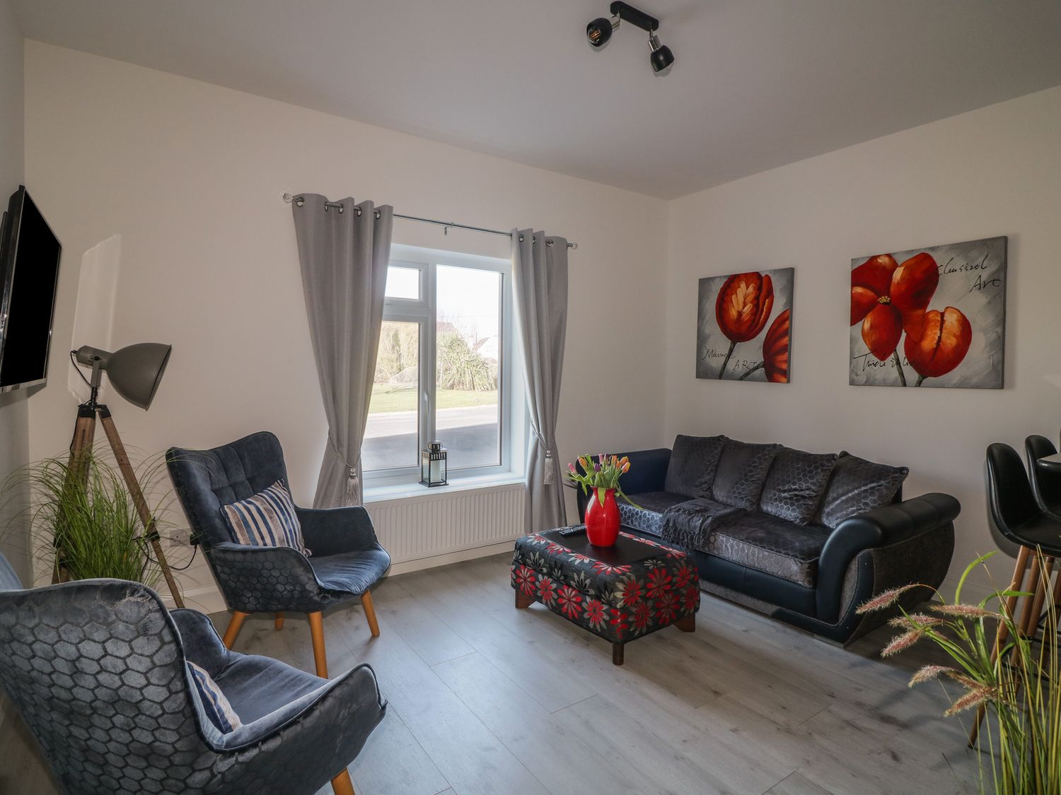 A living room with a black sofa two blue chairs a window with grey curtains and floral paintings at Beach Row in Fethard-On-Sea