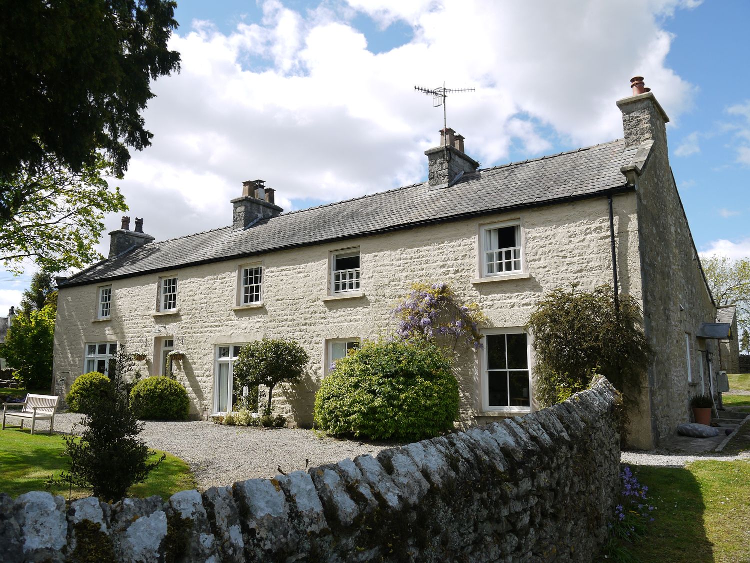 A stone cottage with multiple chimneys surrounded by bushes and a stone fence at 1 The Green in Silverdale