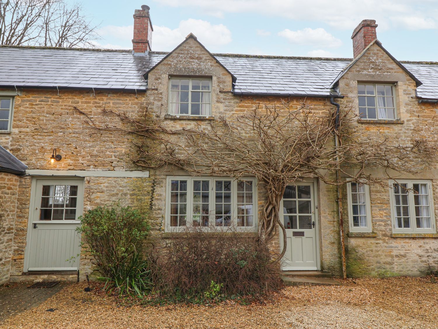 A stone cottage with multiple windows a vine climbing around the door and bushes in front at Little Lemhill Farm Cottage in Lechlade-On-Thames