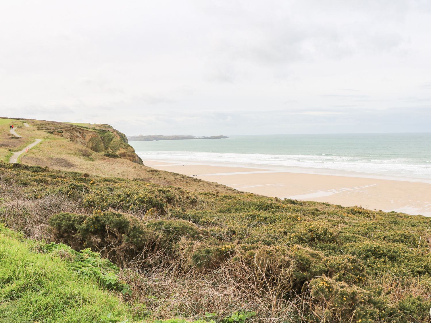 A sandy beach with waves and a grassy cliffside with a walking path at Coastmans Nest in Mawgan Porth