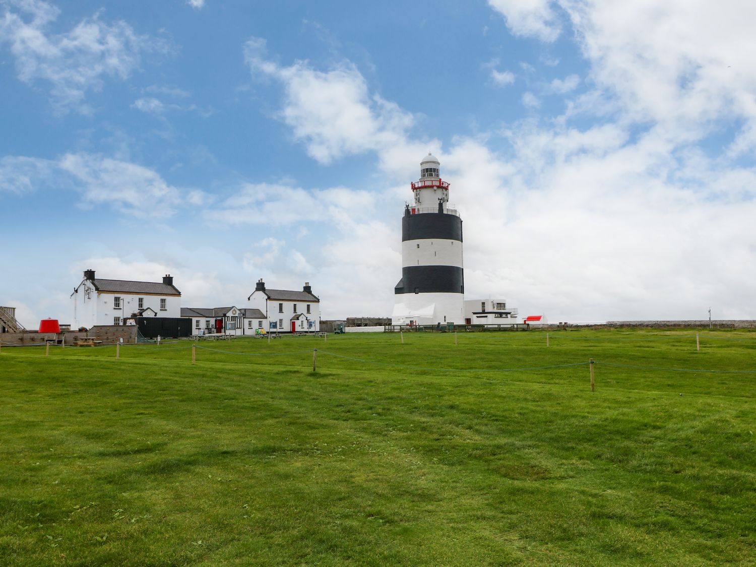 A lighthouse with black and white stripes next to white buildings on grass with a blue sky at The Dairy in Clonsharragh near Duncannon County Wexford