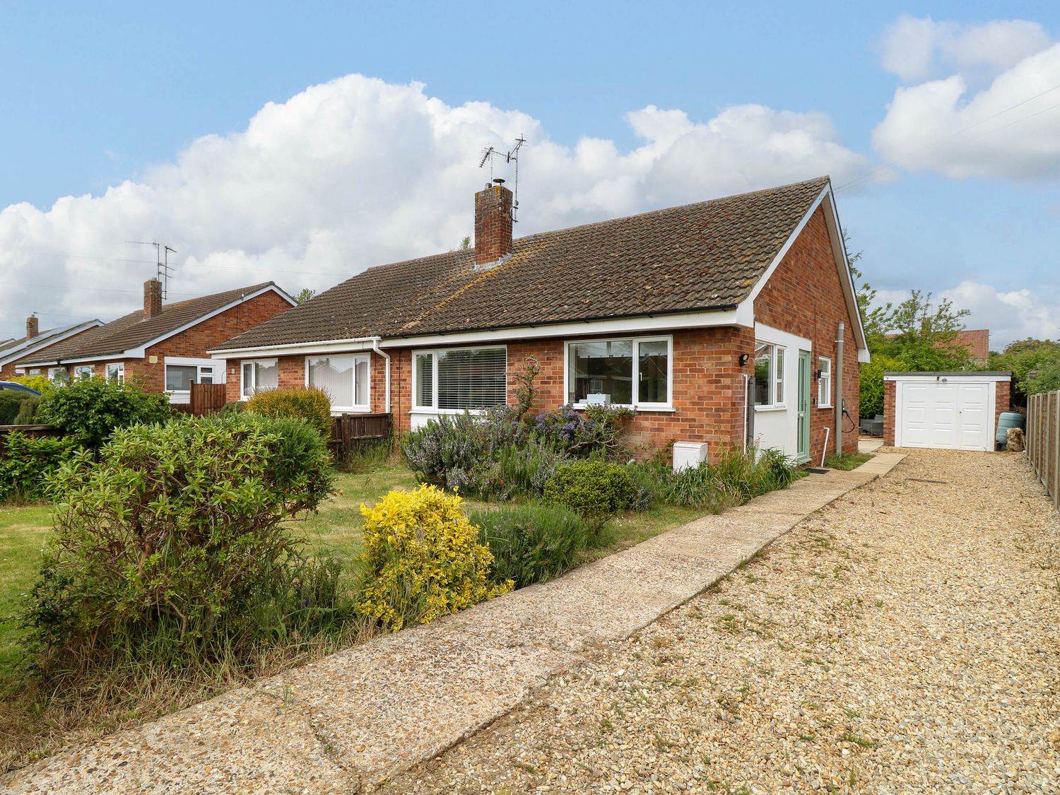 A single-story brick house with a tiled roof a front garden with shrubs and a gravel driveway leading to a garage at Little Lodge in Dersingham