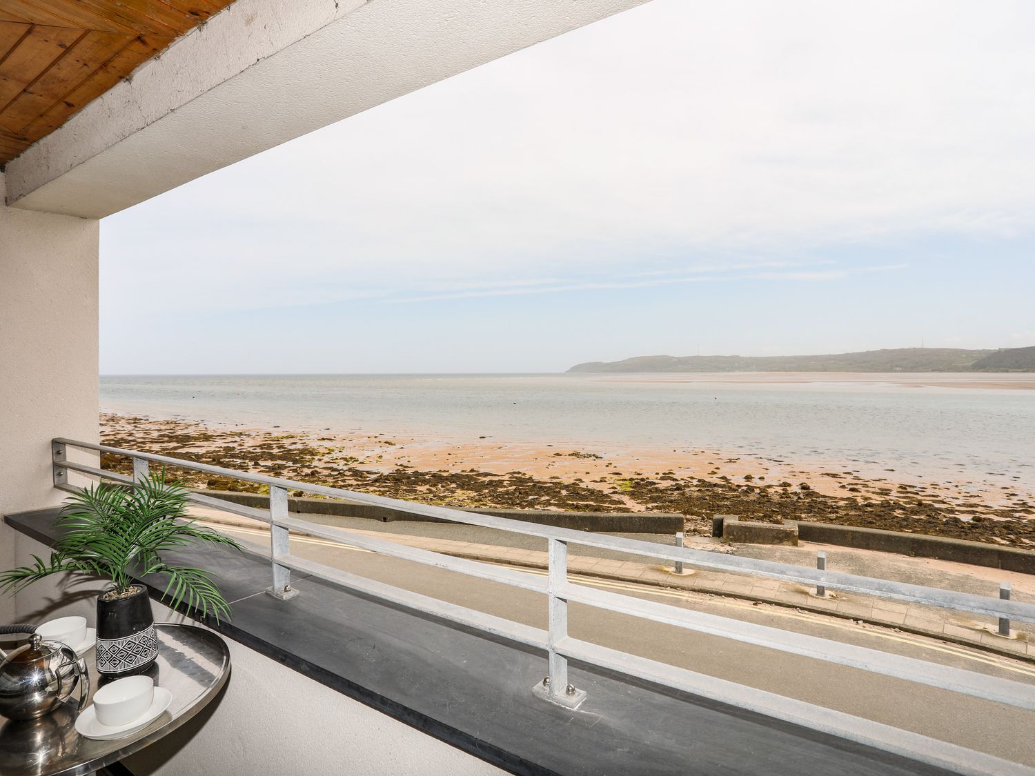 A balcony with a table holding a plant and tea set overlooking a beach and sea at 2 The Quay in Pentraeth