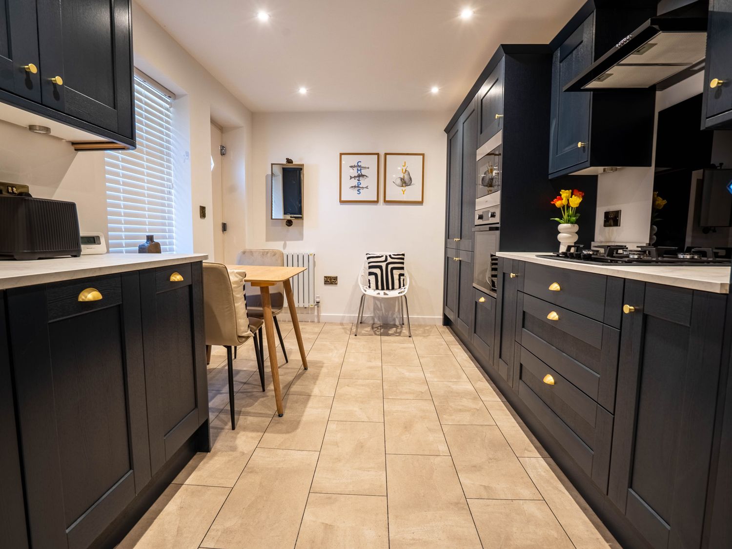 A kitchen with dark cabinets a wooden table with chairs two framed pictures and tiled floor at The Cove in Kessingland
