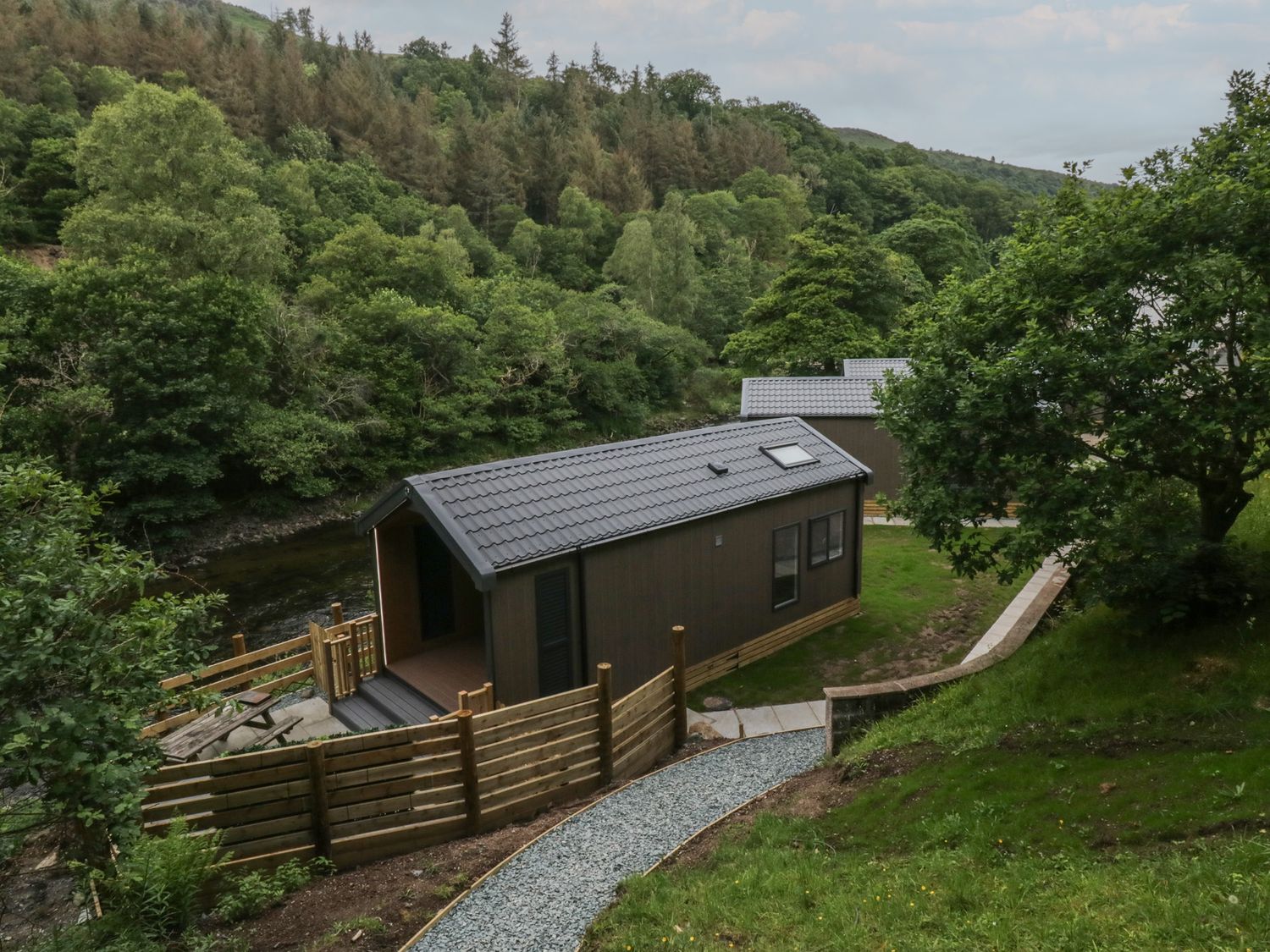 Two cabins near a river with trees in the background at Dubs Luxury Family Pod in Keswick