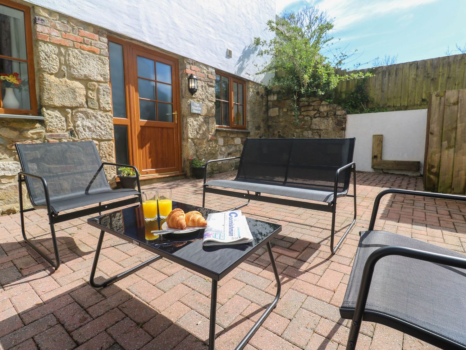 An outdoor patio with black metal chairs and a table with croissants orange juice and a newspaper at The Stable Barn in Hayle