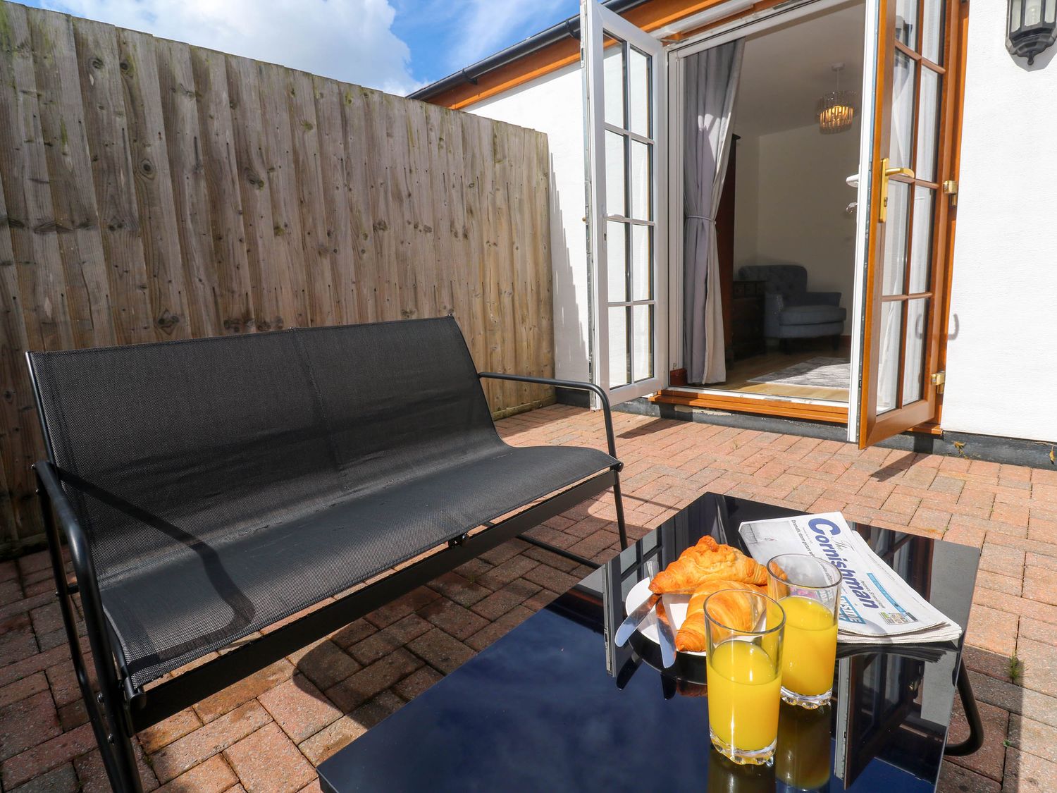 An outdoor patio with a black bench and a table with croissants orange juice and a newspaper near an open door showing an indoor chair and curtain at The Baler House in Hayle