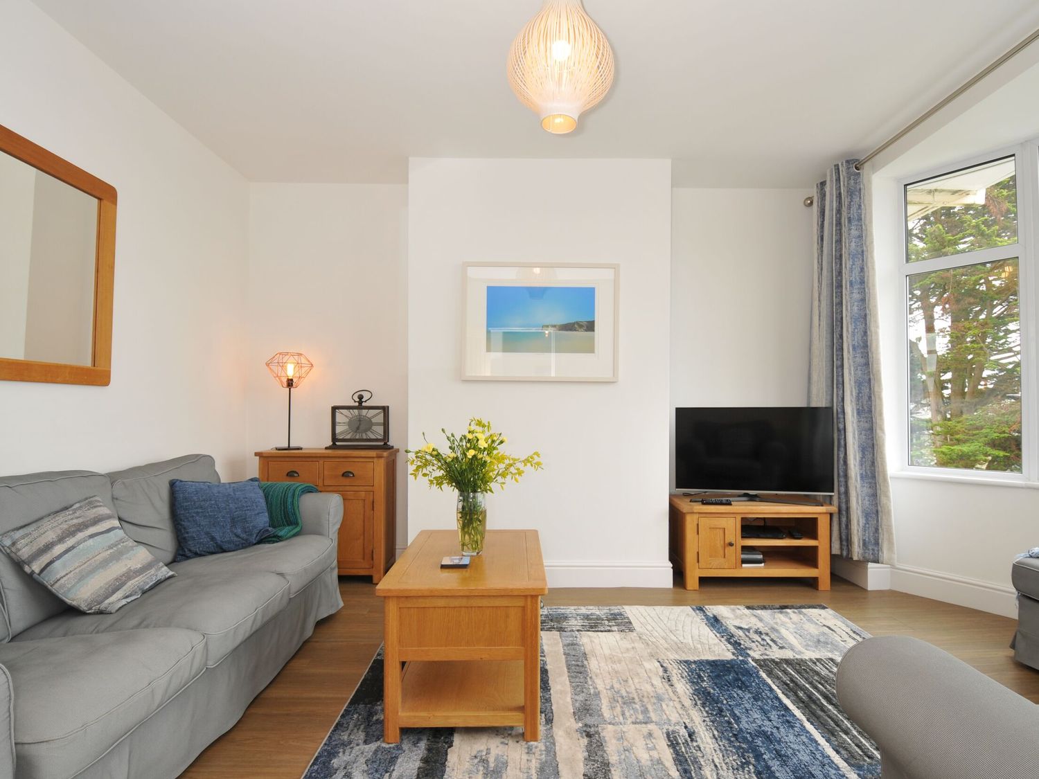 A living room with a gray sofa a wooden coffee table with flowers a wooden cabinet with a lamp and a television on a wooden stand at Falmouth Bay 2 in Falmouth