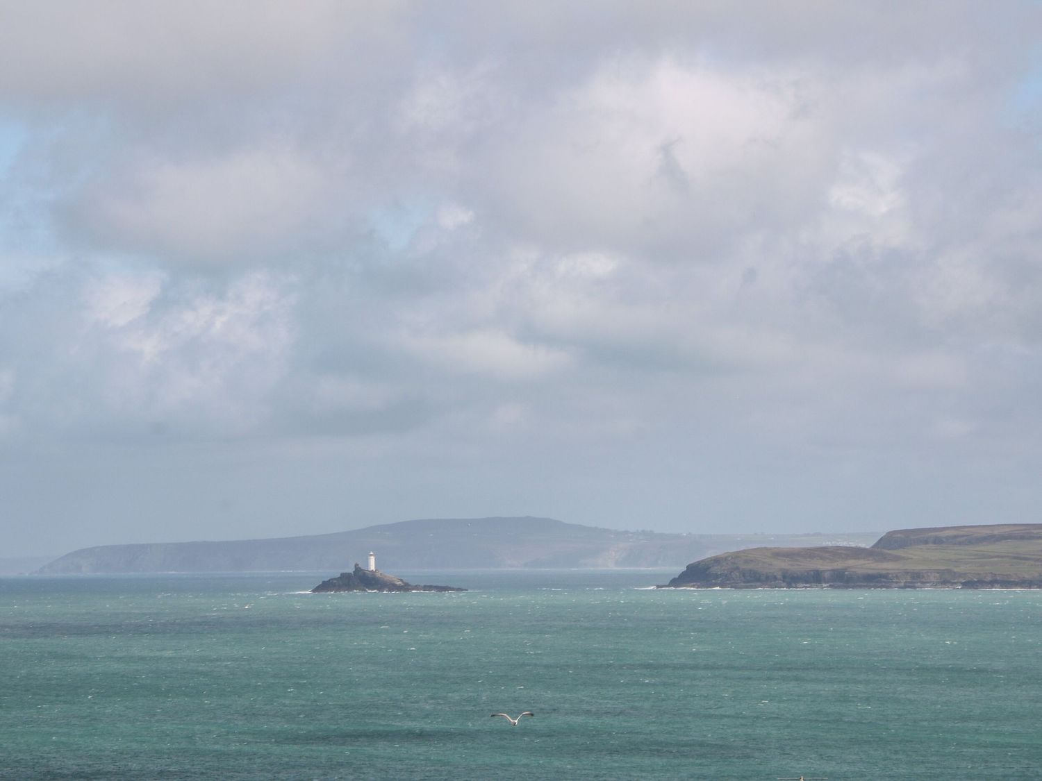 A seascape showing a small rocky island with a lighthouse and distant land in the background at Chy Benyngoth in Hayle