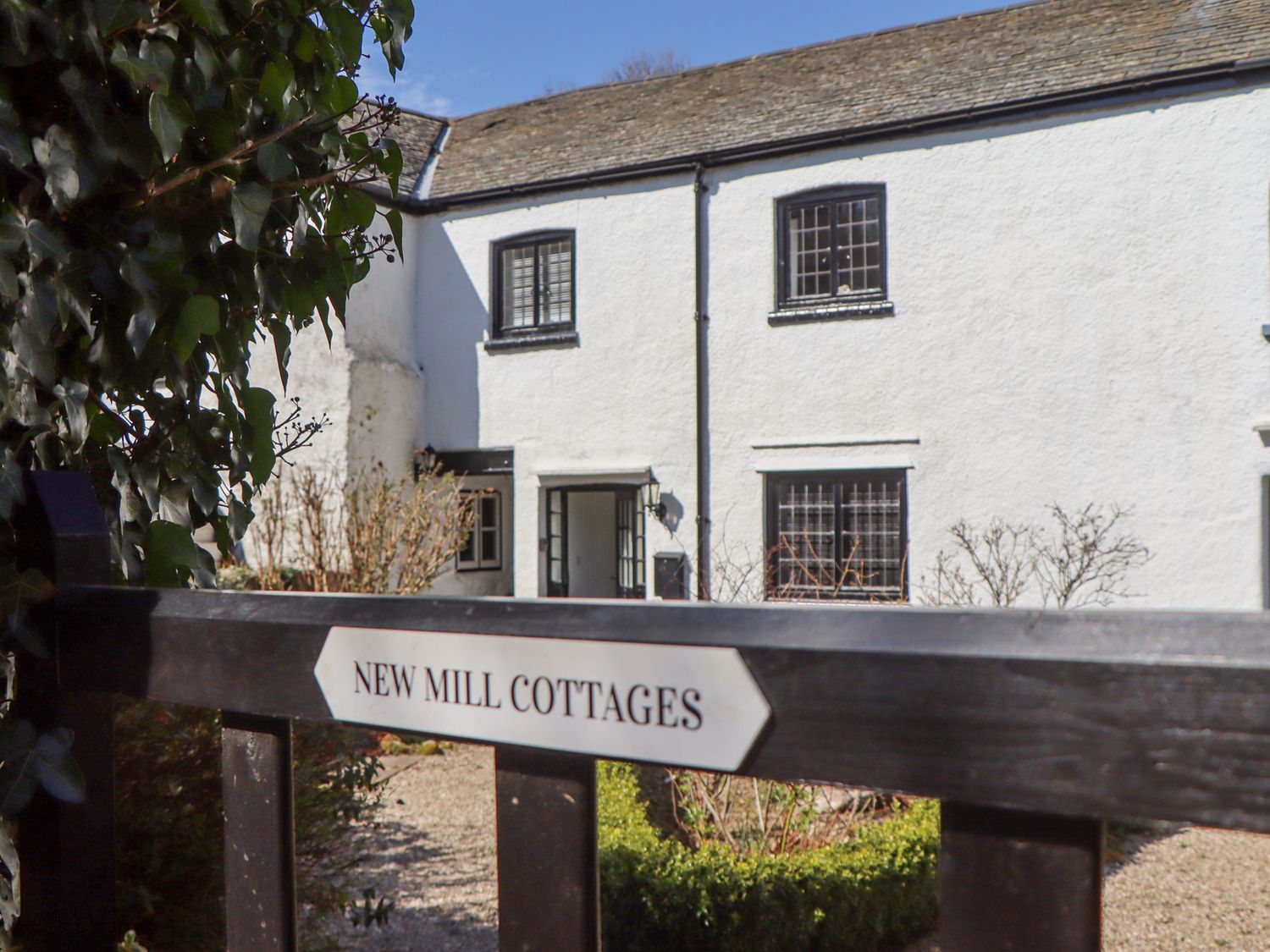 A white cottage with black-framed windows and an open door behind a gate with New Mill Cottages sign at 2 New Mill Cottages in North Bovey