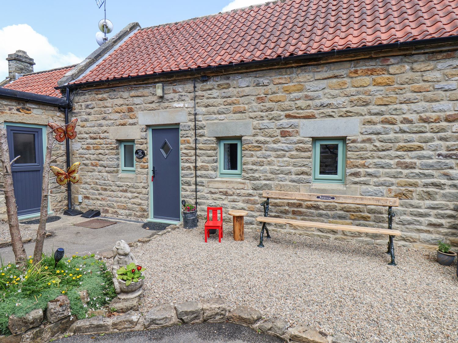 An exterior stone wall of a building with a blue door and windows a wooden bench a small wooden stool a red child size chair and garden decorations at Wayside Farm Dairy Cottage Cloughton near Burniston