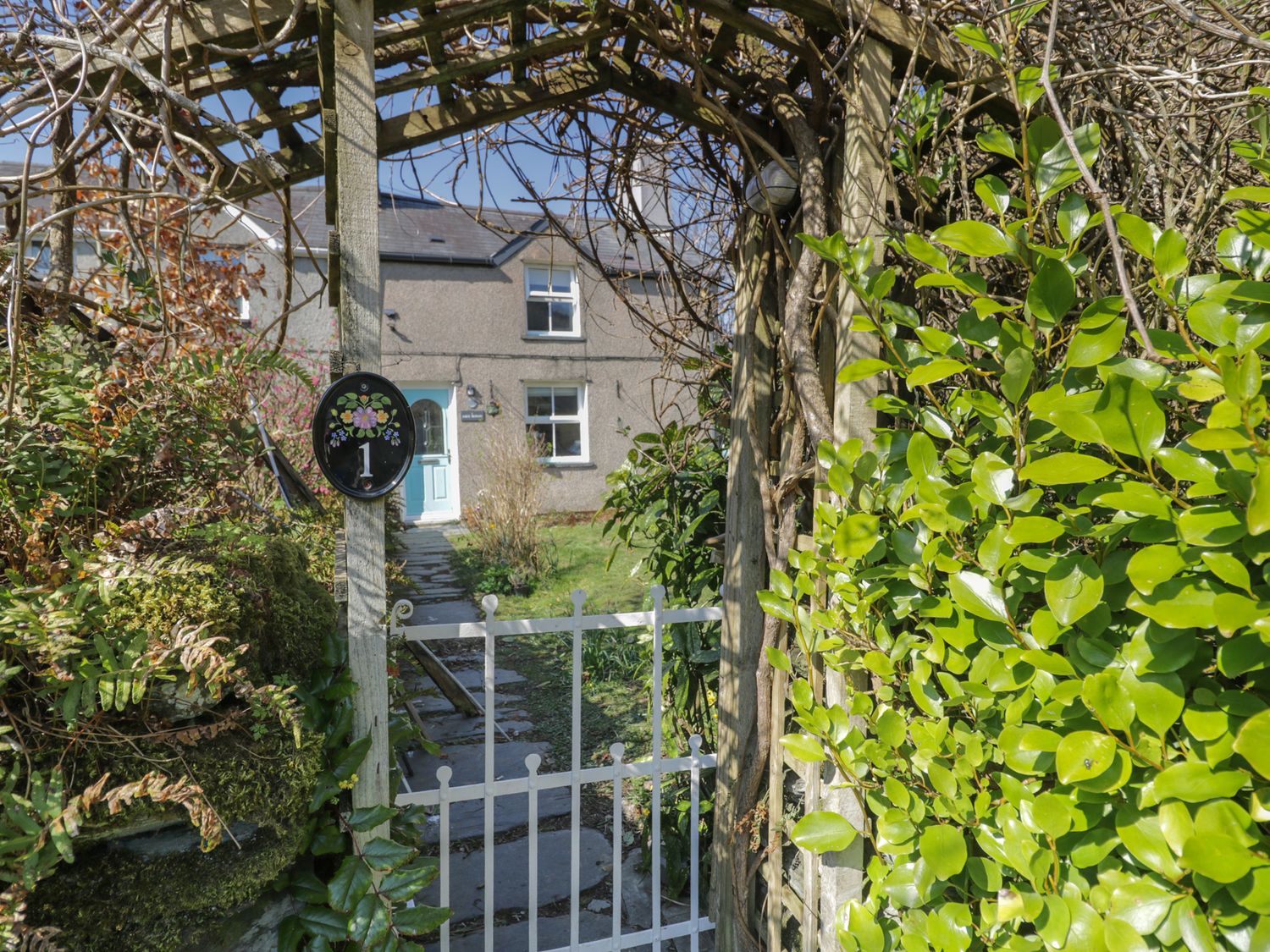 A pathway with stone slabs leading to a house with a blue door seen through a wooden arch with a white gate and foliage at 1 Aberbeinw in Dolwyddelan