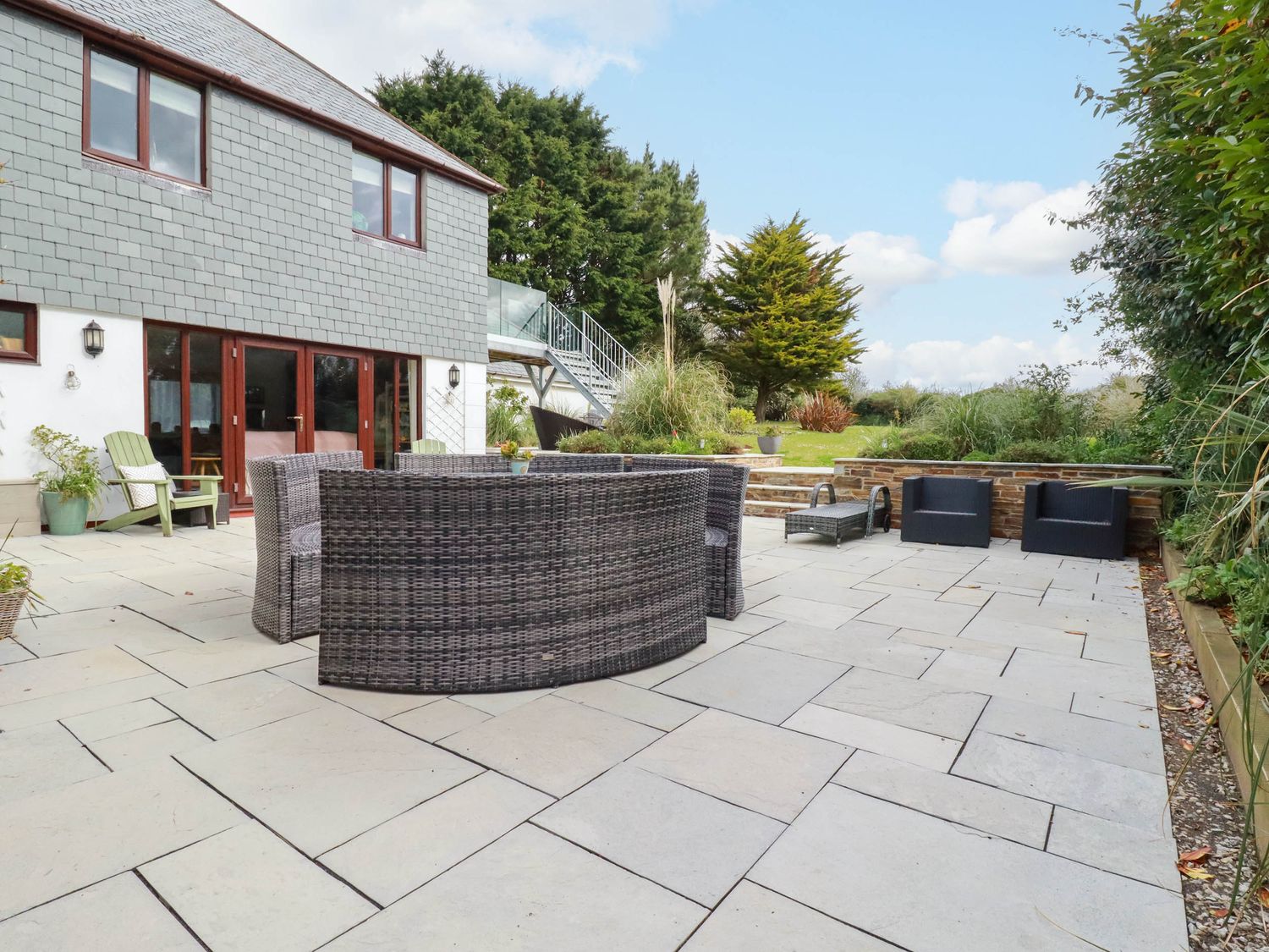 A paved patio with outdoor furniture and a garden with trees behind at Goatsland in Trebetherick