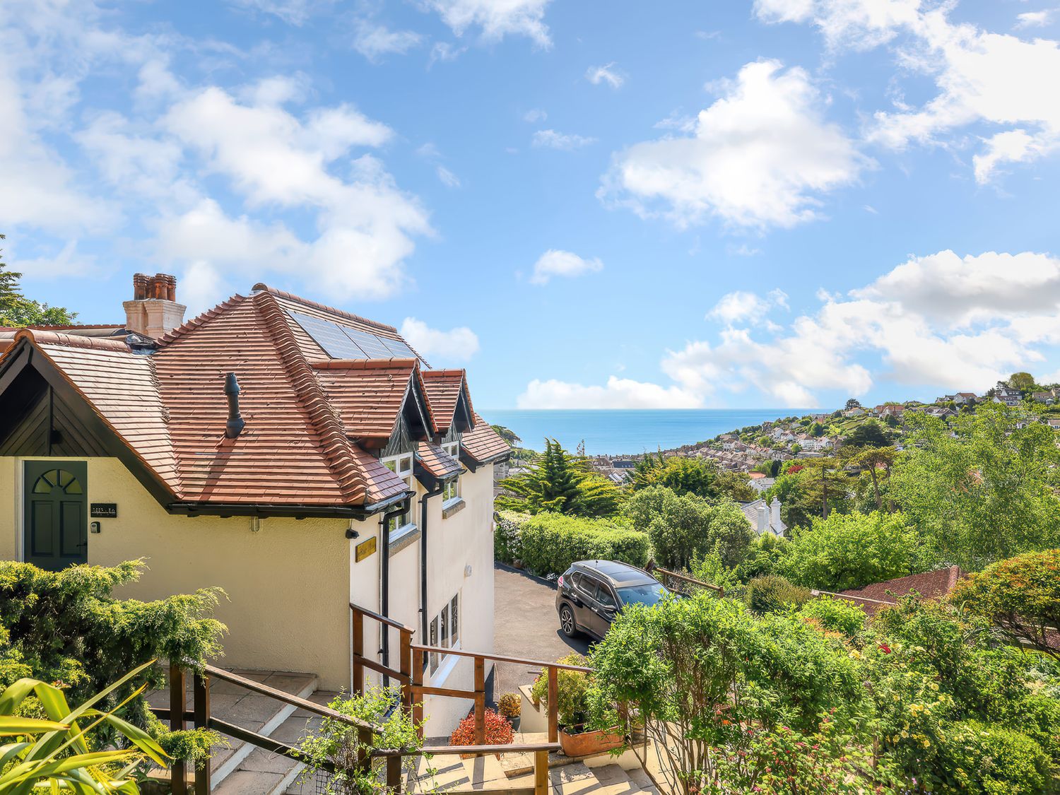A house with a red tiled roof and solar panels surrounded by greenery with a black car parked and a sea view in Beer