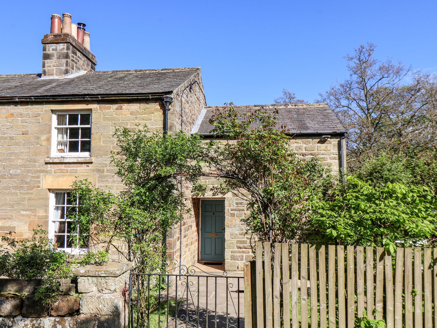 A stone cottage with a blue door surrounded by greenery and a metal gate at Whinstone Cottage Esk Valley near Grosmont