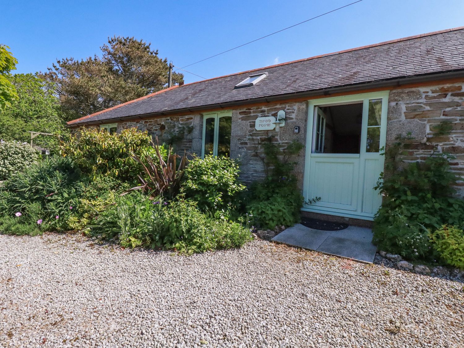 A stone building with green doors and windows surrounded by plants and gravel at The Old Piggery in Porthtowan