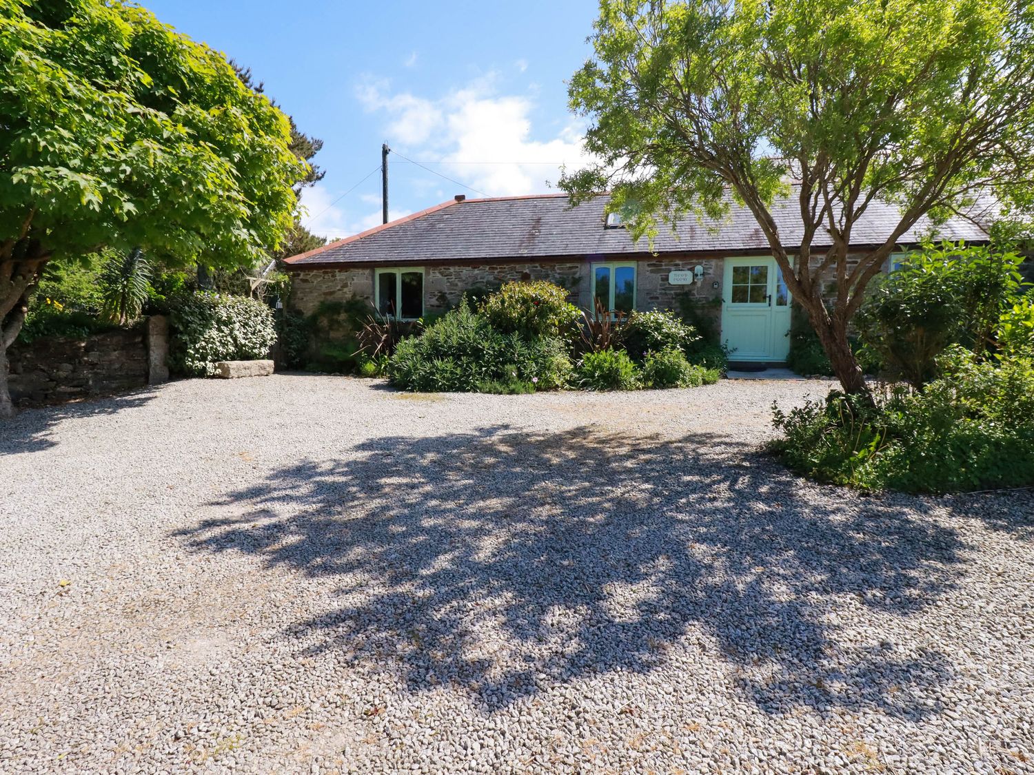 A stone building with a green door and windows surrounded by trees and gravel driveway at The Old Piggery in Porthtowan