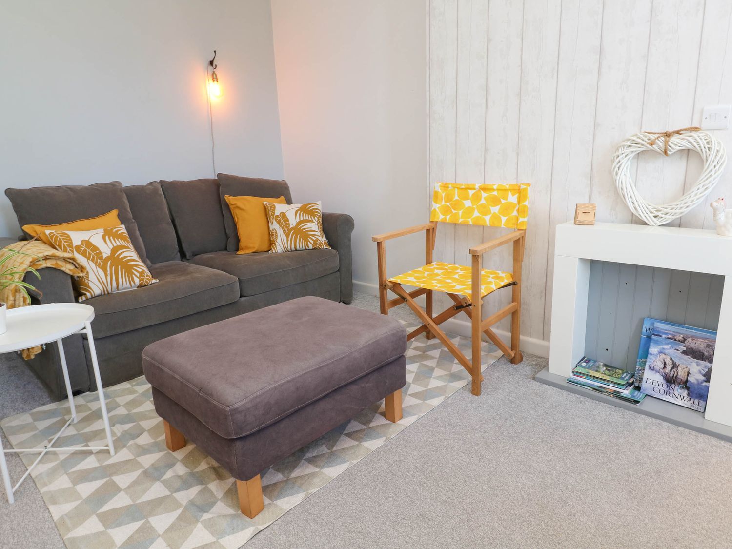 A living area with a grey couch with yellow cushions a wooden chair with yellow fabric a grey ottoman and a white small table at Penzance Apartment in Penzance