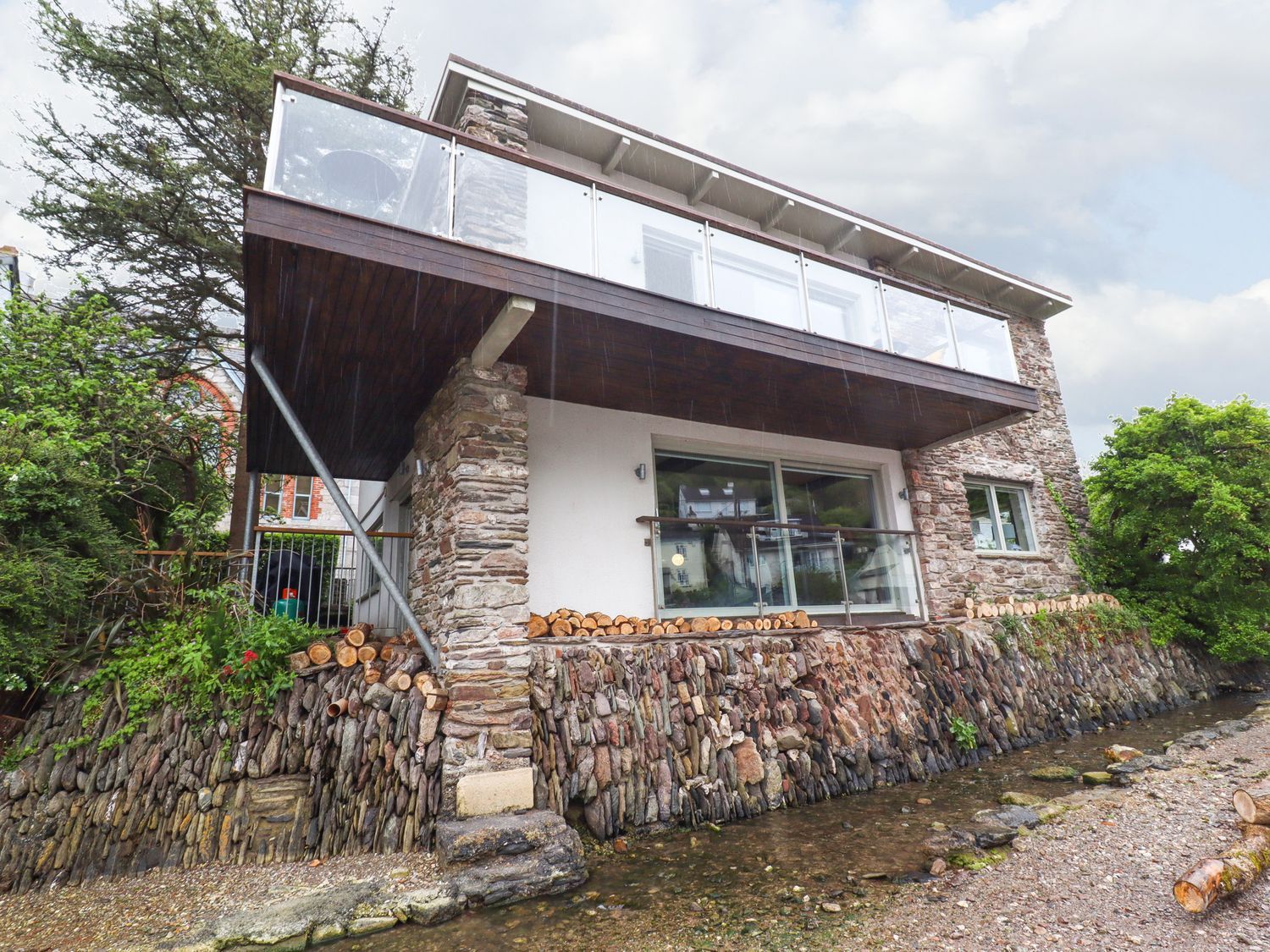A stone house with a balcony and glass railing by a river at Tidal Waters in Noss Mayo