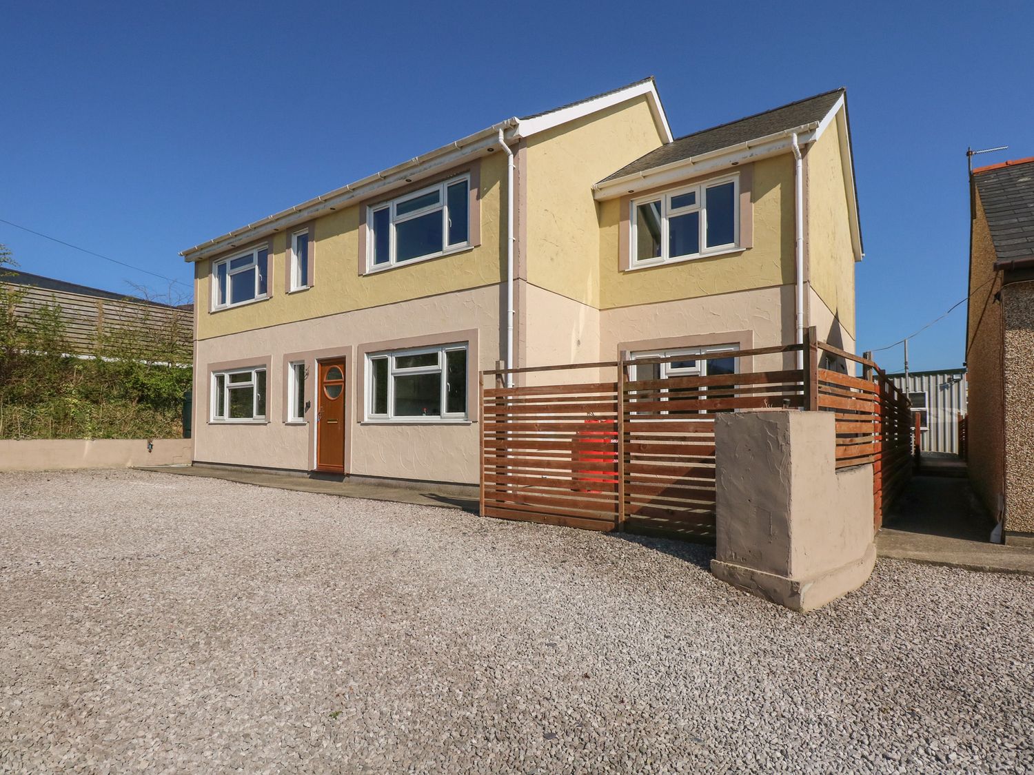 A two-story house with yellow and beige walls with a brown wooden door and multiple windows with a gravel driveway and a wooden fenced area at Criw Newydd in Bryncroes near Sarn Meyllteyrn