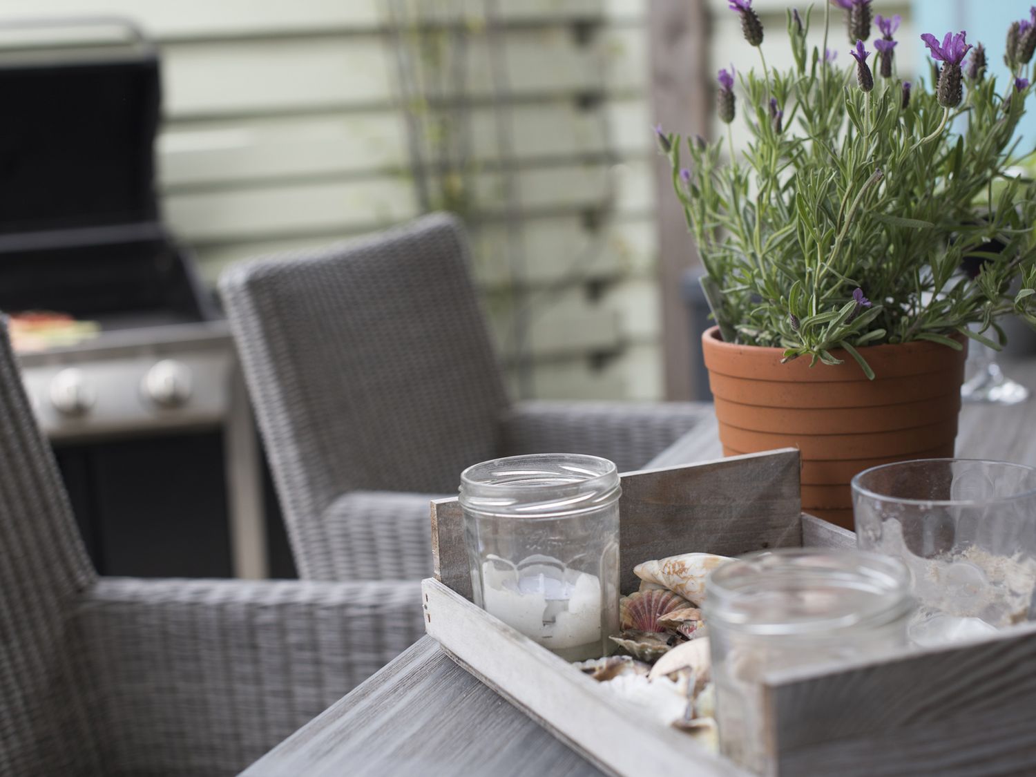 A patio table with a wooden tray holding glass jars and seashells next to a potted lavender plant with a barbecue and wicker chairs in the background at Two Bare Feet Rose near Perranporth