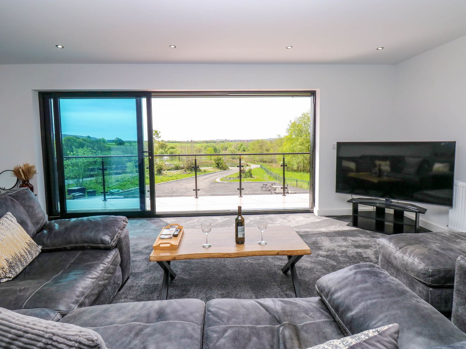 A living room with grey sofas a wooden coffee table a large flat screen tv and sliding glass doors overlooking a green landscape at Sycamore Cottage in Frizington