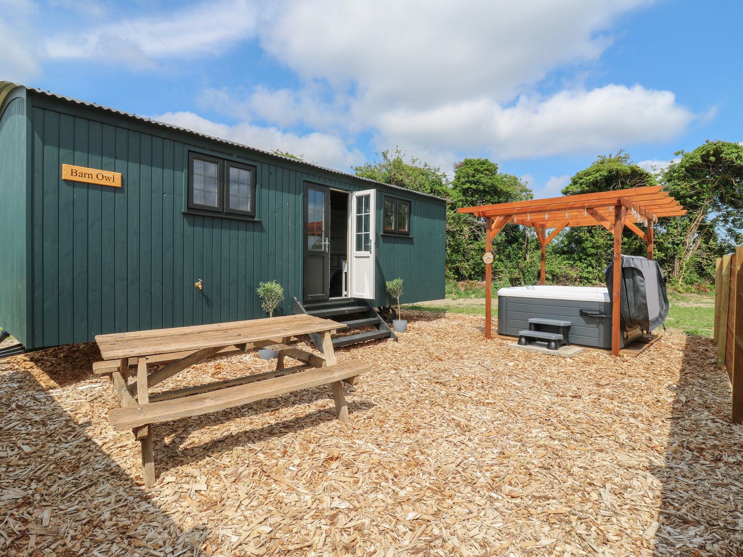 An outdoor area with a green wooden cabin labeled Barn Owl a wooden picnic table and a hot tub under a wooden pergola at Barn Owl in Lode near Bottisham