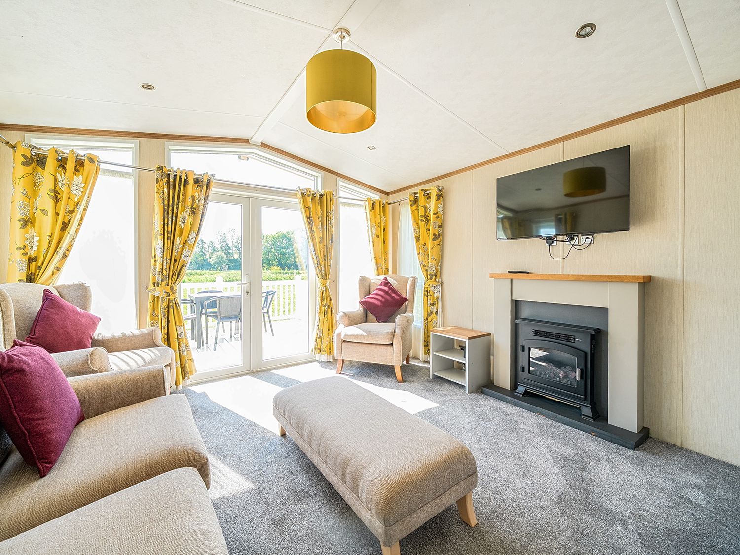 A living room with beige armchairs and a sofa with purple cushions a gray ottoman a fireplace with a television above yellow floral curtains and a table with chairs outside Serenity Shores -  Billing Aquadrome in Billing Aquadrome Holiday Park