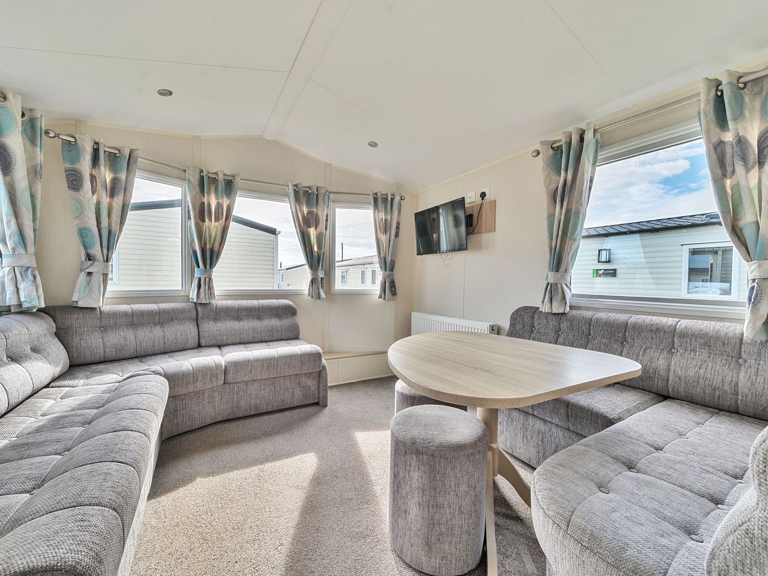 A living room with grey upholstered seating a wooden table and multiple windows with patterned curtains at The Lookout - Hayling Island in Hayling Island Holiday Park